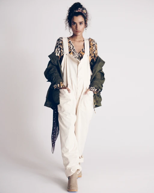 Full-shot of a woman in white overalls, an animal-print top, and a green jacket, standing in a photo studio with a white backdrop.