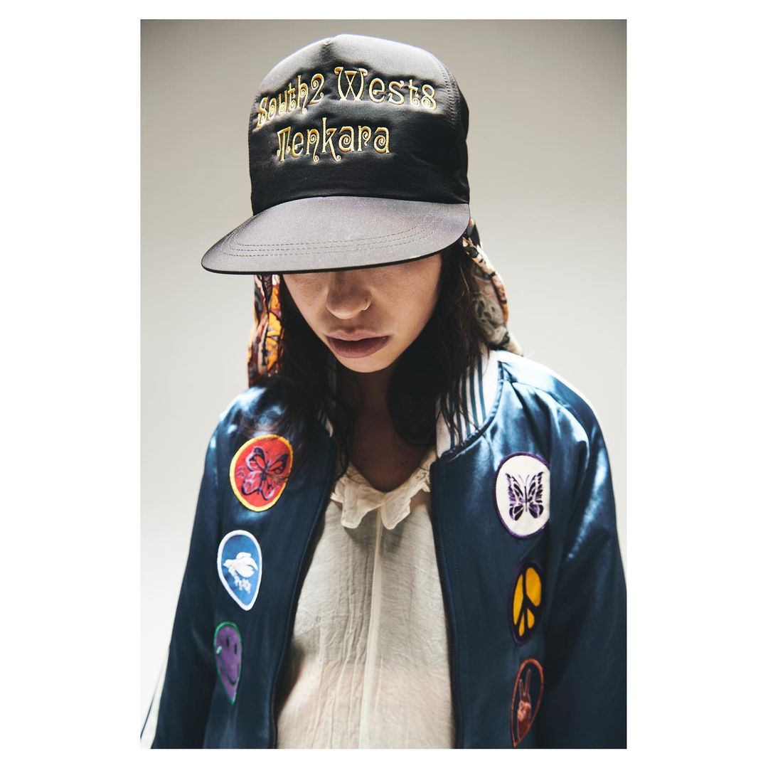 Woman wearing a cap and patched satin jacket in a studio shot. Model with dark hair poses looking down in fashion style studio lighting.