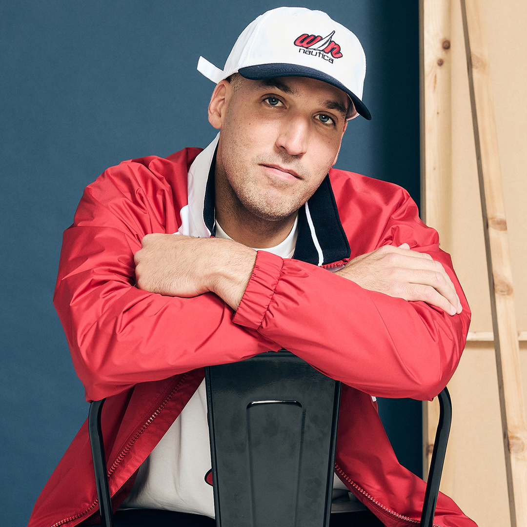 Adult male with cap seated on black chair wearing a red jacket in a medium shot with a blue and tan background.