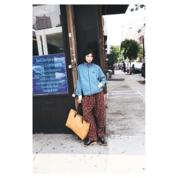Model standing on a New York sidewalk wearing a patterned blue jacket and brown trousers, holding a large yellow tote bag.