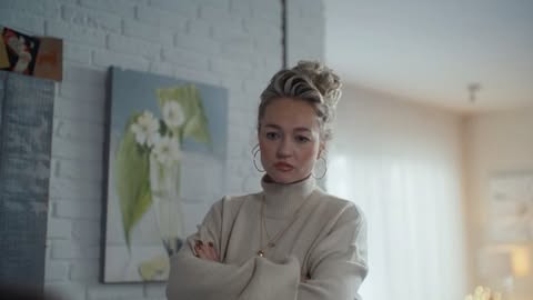 Indoor portrait of a blonde woman in a turtleneck sweater with her arms crossed, standing in front of a painting and brick wall.