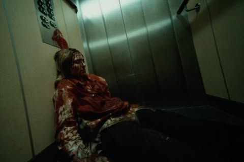 Woman covered in red liquid sits in a metal elevator with control panel and hand rail. The scene is dark with harsh lighting.