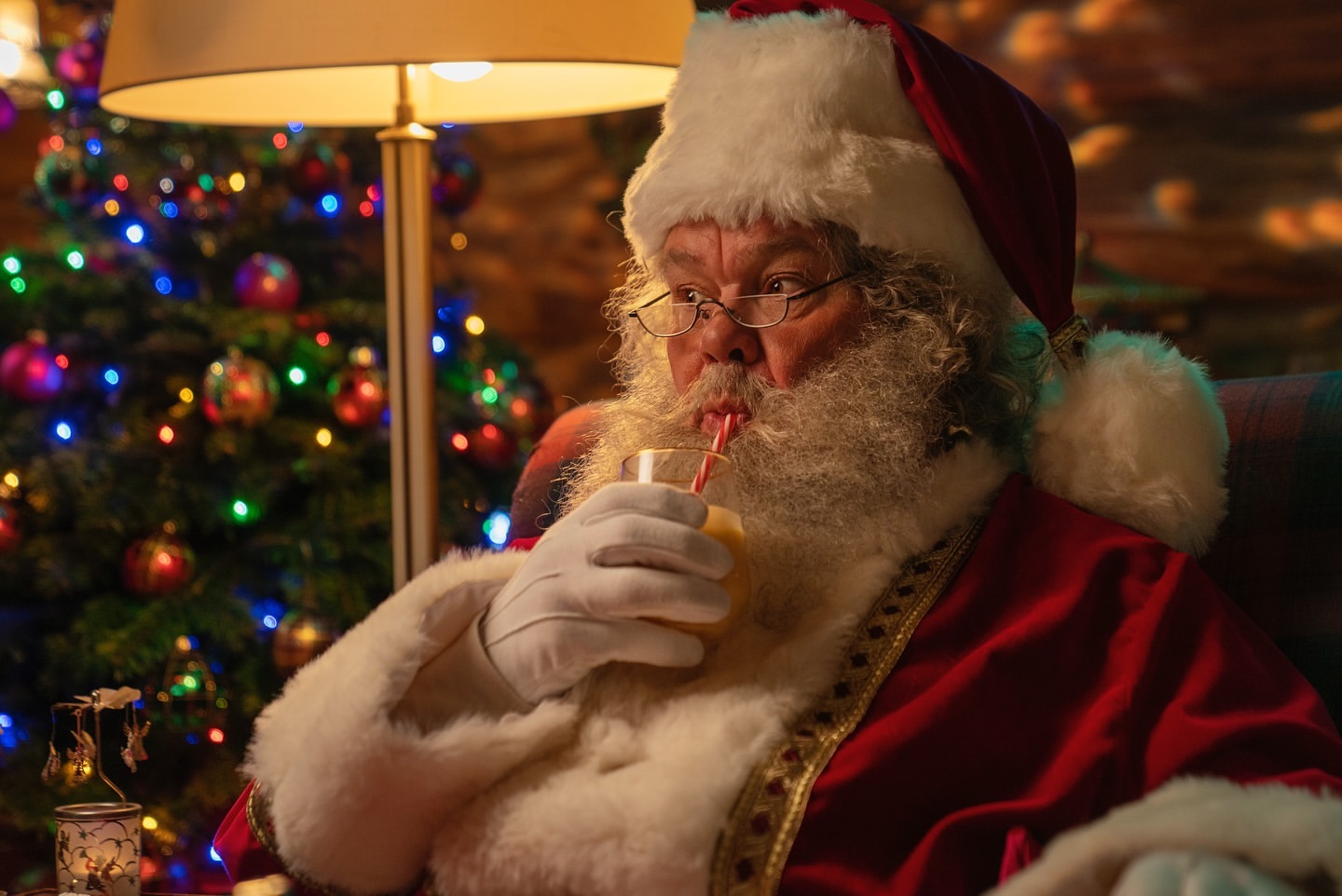 Santa Claus in a red suit drinking from a glass with a straw, sitting indoors near a Christmas tree with colorful lights.