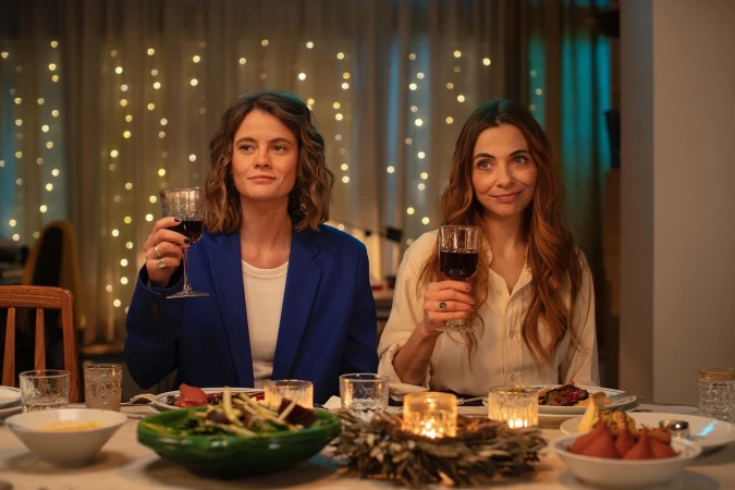 Two adult women at an indoor dinner table raising wine glasses, dressed in casual-chic attire, celebrating against a soft bokeh light backdrop.