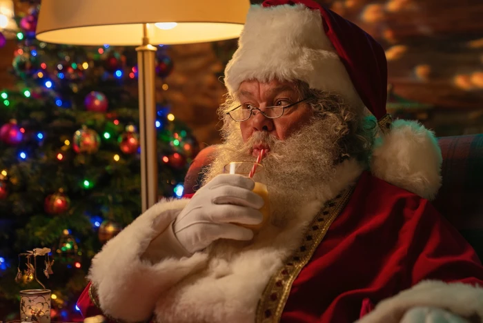Santa Claus in a red suit and white gloves drinking from a glass with a straw indoors near a decorated Christmas tree.