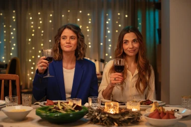 Two women holding glasses of red wine seated at a dining table with string lights in the background