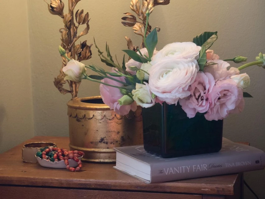 Indoor still life showcases a floral arrangement with pink and white flowers in a black vase, resting on a book atop a wooden table.
