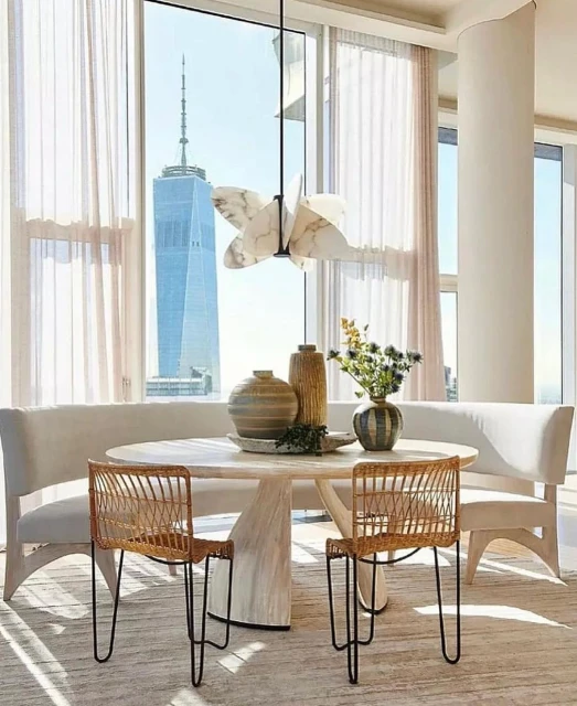 Bright, airy dining room featuring a round table with wicker chairs, a cushioned bench, and a large window with a city view.
