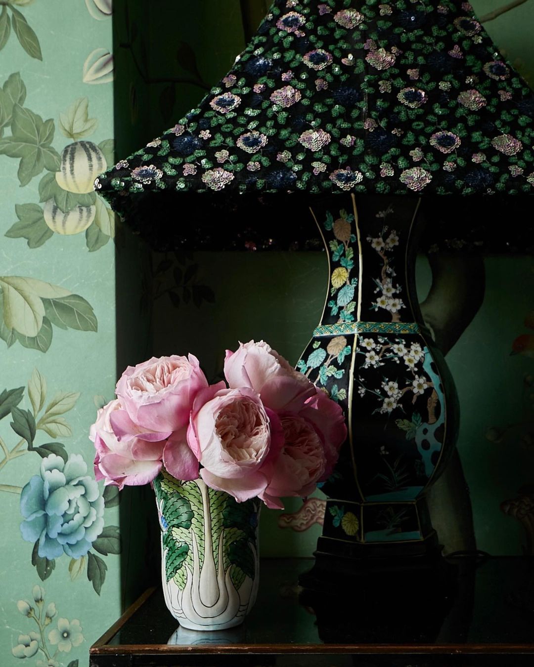 Table lamp with a black sequin lampshade and a floral vase with pink flowers sits on a black surface against a floral wallpaper.