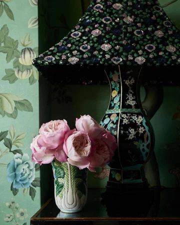 Table lamp with a black sequin lampshade and a floral vase with pink flowers sits on a black surface against a floral wallpaper.