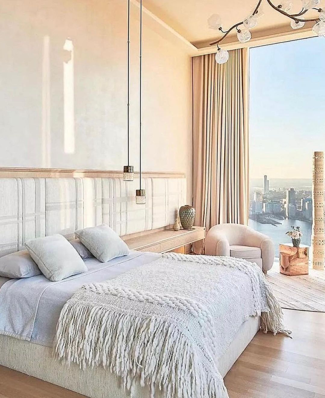 Bedroom interior with a large bed featuring white pillows and a fringed blanket, pendant lights, an armchair, and a city view.