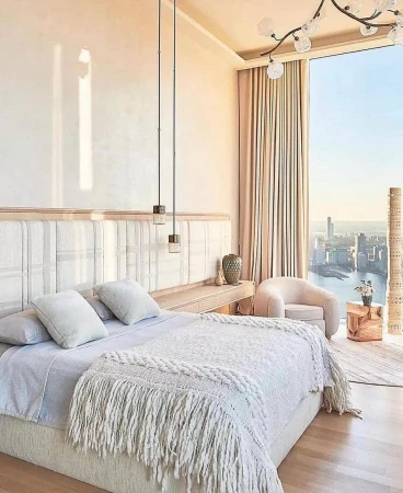 Bedroom interior with a large bed featuring white pillows and a fringed blanket, pendant lights, an armchair, and a city view.