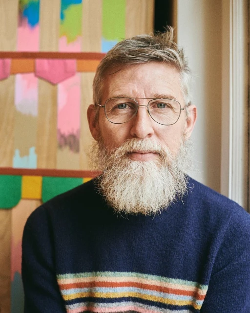 Close-up portrait of a mature man with a grey beard, wearing glasses and a striped sweater, posing in front of colourful artwork.
