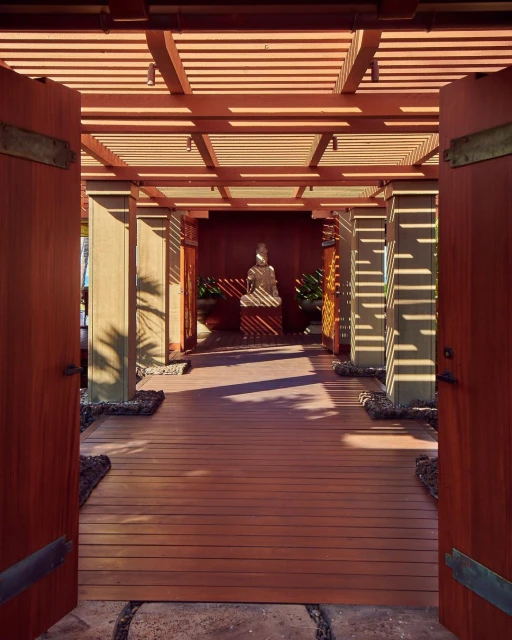 Wood walkway leads to Buddha statue under pergola, light and shadow interplay create a tranquil ambiance in architectural space.