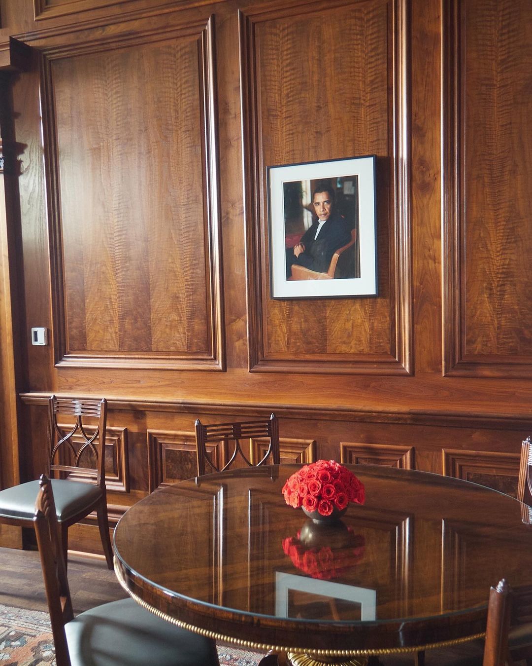 Formal dining room featuring a glossy round table with chairs, an arrangement of red roses, and a portrait on the wood-paneled wall.