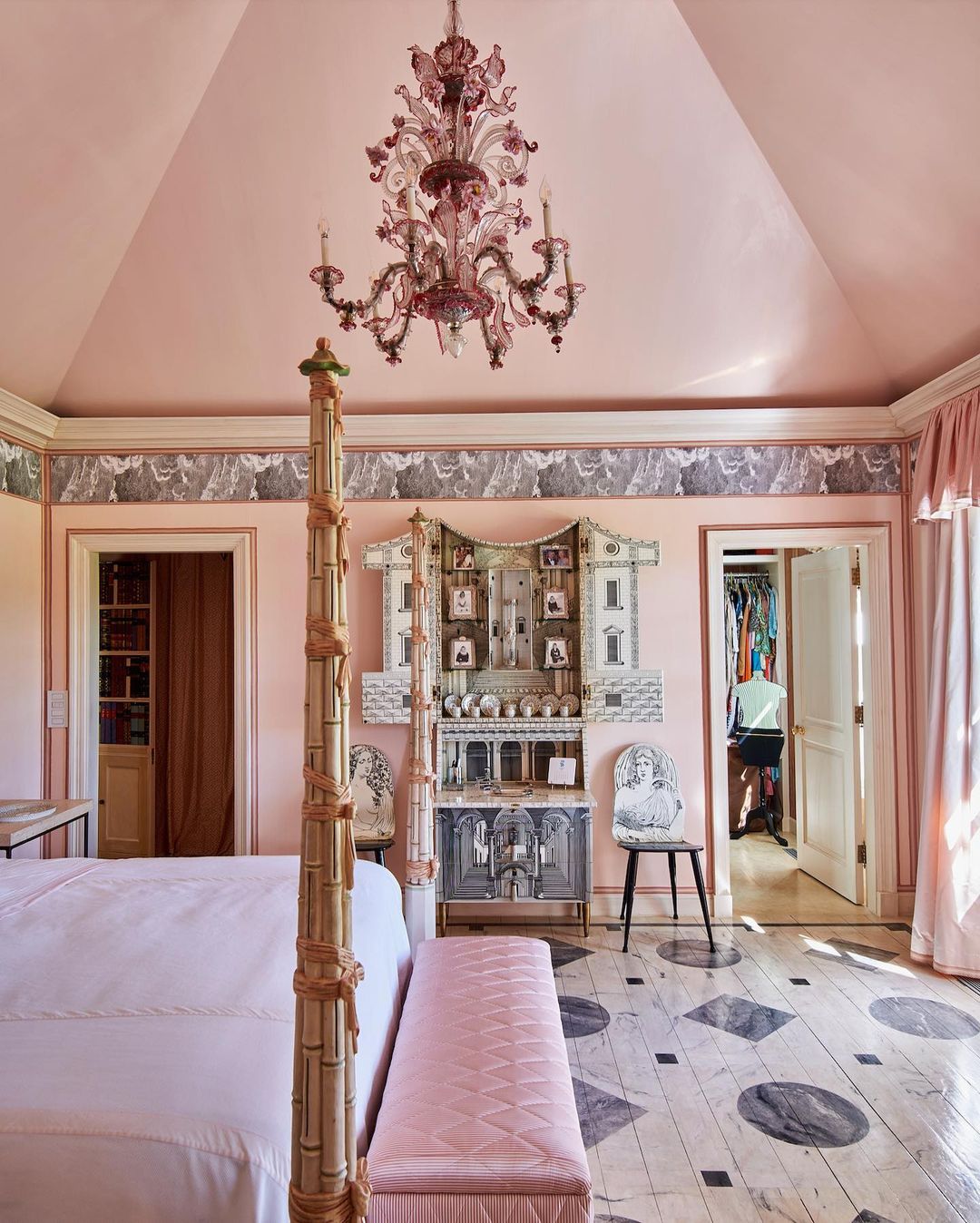 Bedroom interior featuring a four-poster bed, pink walls, decorative chandelier, ornate cabinet, and a person in the background.