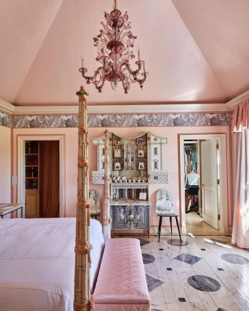 Bedroom interior featuring a four-poster bed, pink walls, decorative chandelier, ornate cabinet, and a person in the background.