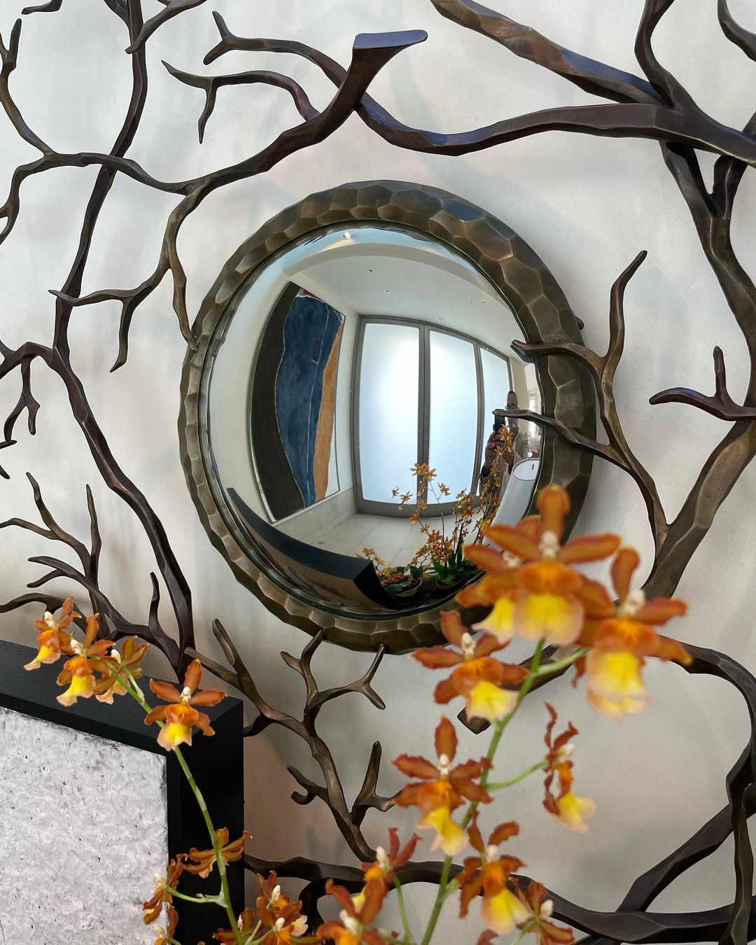 Round mirror with hammered metal frame surrounded by metal tree branches and orange orchids in an indoor environment. Interior design.