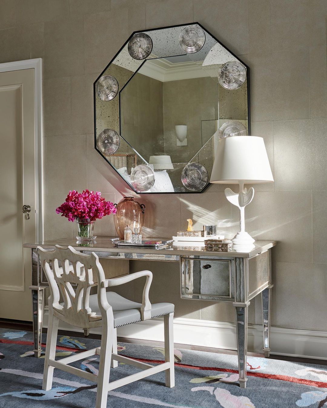 Home interior featuring a mirrored writing desk with pink flowers, lamp, and octagonal mirror in a naturally lit neutral colour scheme.