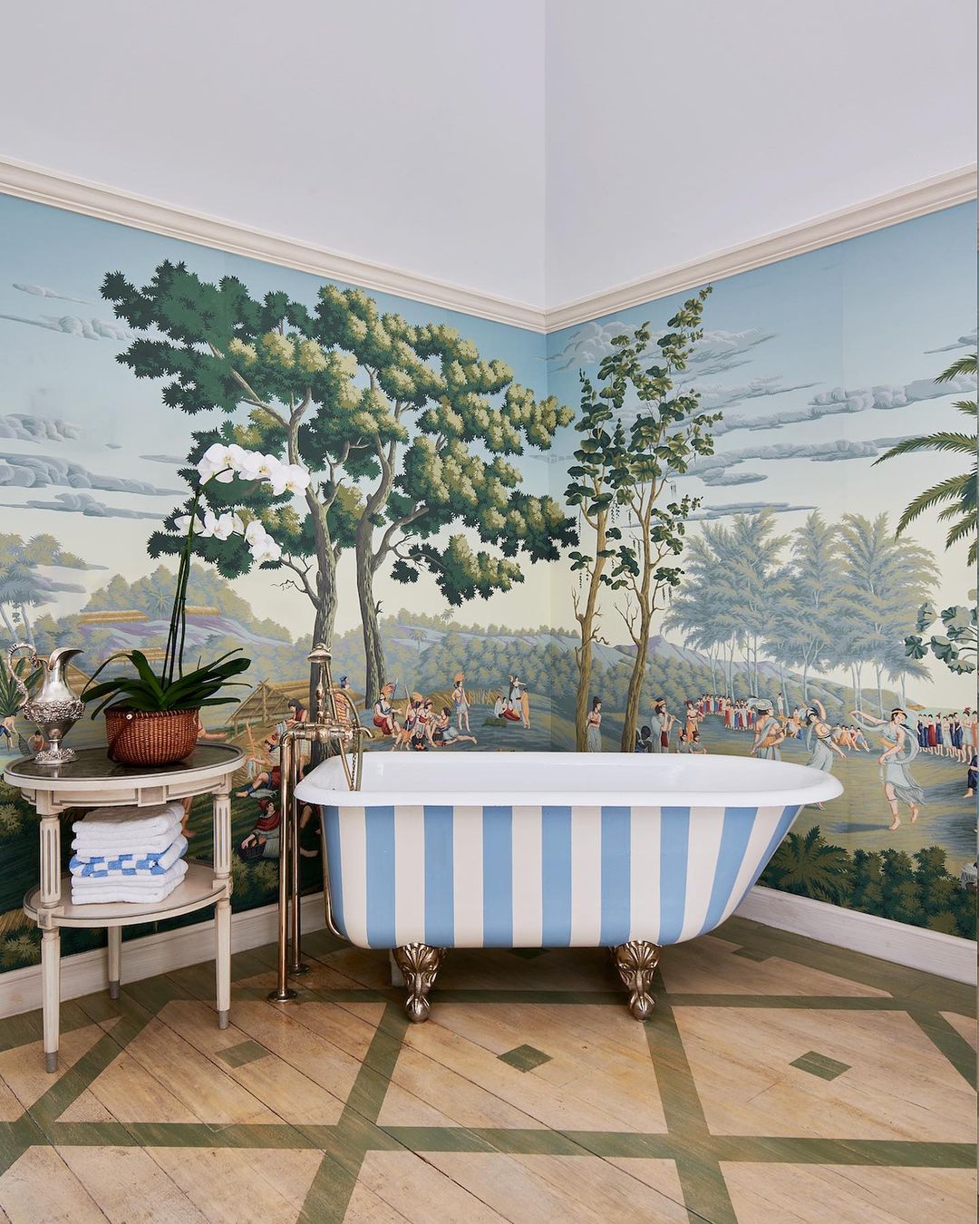 Bathroom interior with blue and white striped bathtub on a wood floor, set against a toile wallpaper with a pastoral scene.