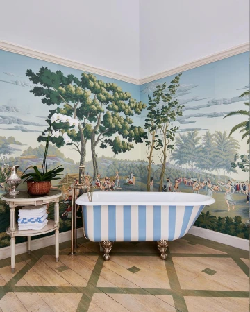 Bathroom interior with blue and white striped bathtub on a wood floor, set against a toile wallpaper with a pastoral scene.