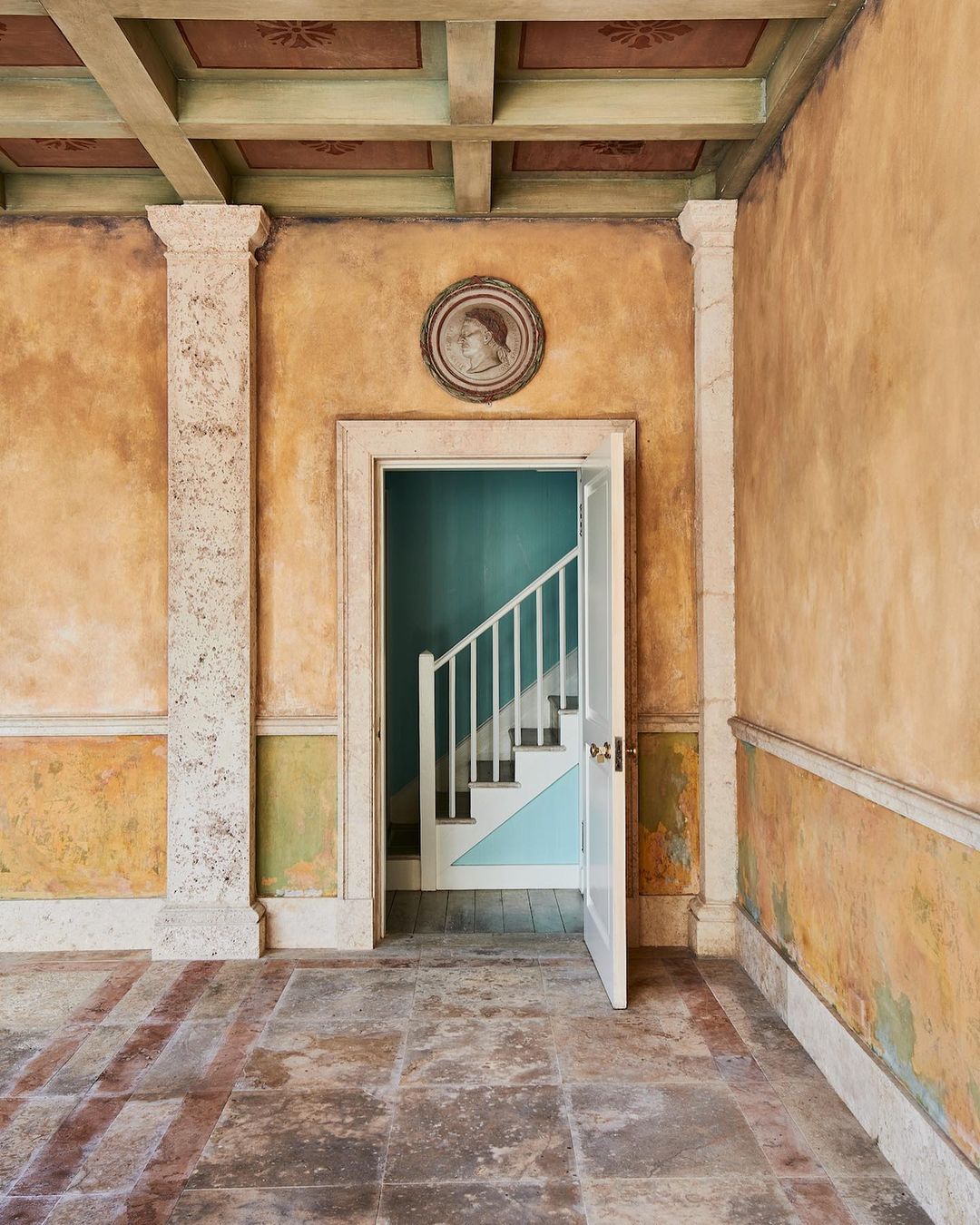 Classical interior featuring ornate plaster walls, coffered ceiling, stone floors, and a view into a stairwell through an open doorway.