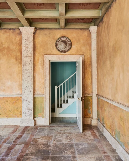 Classical interior featuring ornate plaster walls, coffered ceiling, stone floors, and a view into a stairwell through an open doorway.