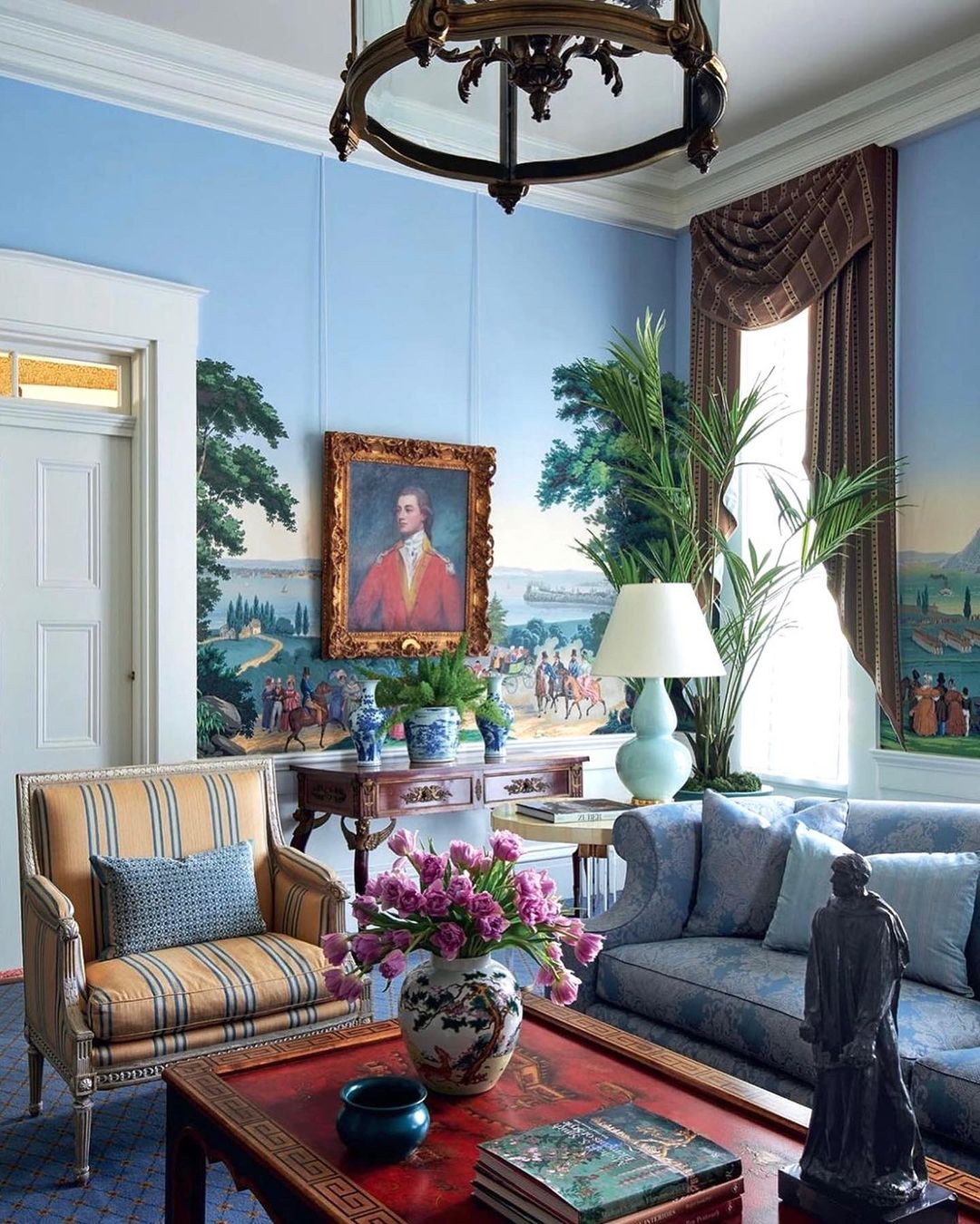 Living room interior featuring blue walls with landscape mural, striped arm chair, sofa, coffee table, and various decorative objects.