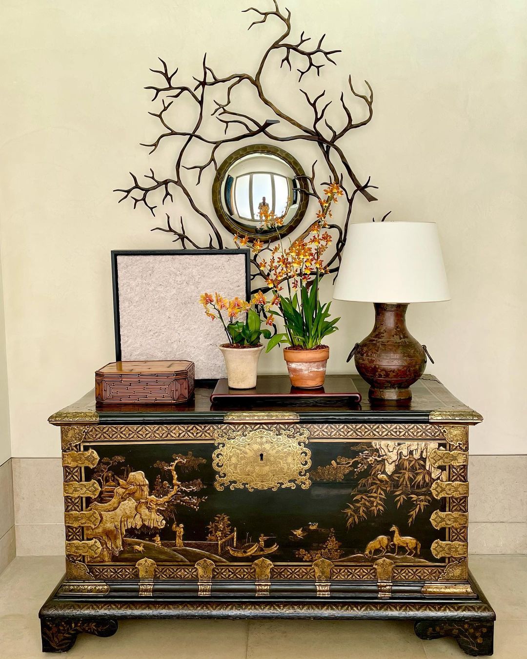 Interior decor with ornate black and gold chest, potted orchids, lamp, and a mirror surrounded by a metal branch sculpture on a cream-colored wall.