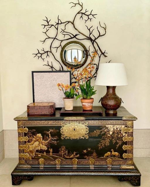 Interior decor with ornate black and gold chest, potted orchids, lamp, and a mirror surrounded by a metal branch sculpture on a cream-colored wall.