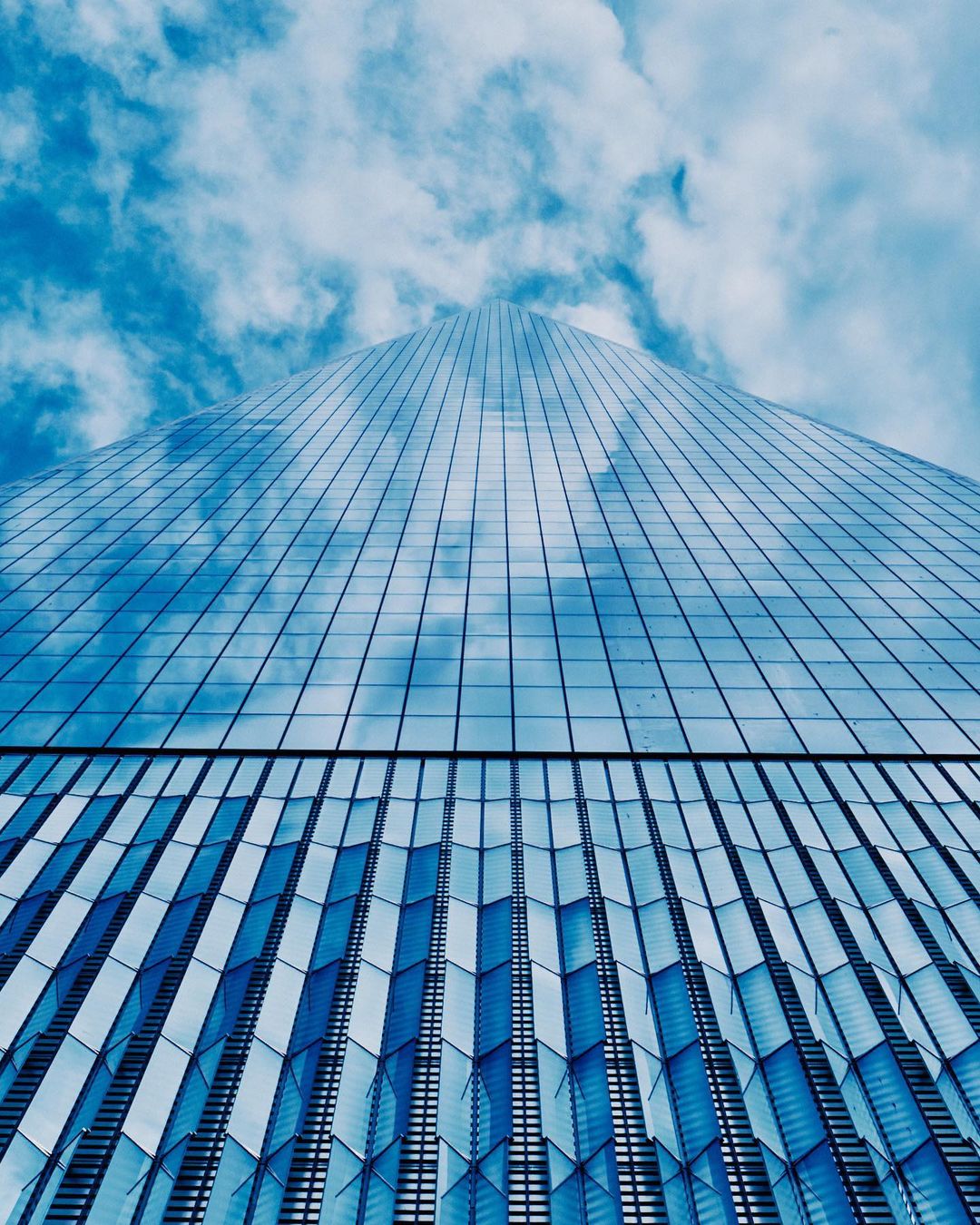 Low angle shot of a modern blue glass skyscraper reaching into a sky with scattered white clouds, emphasizing architectural height.