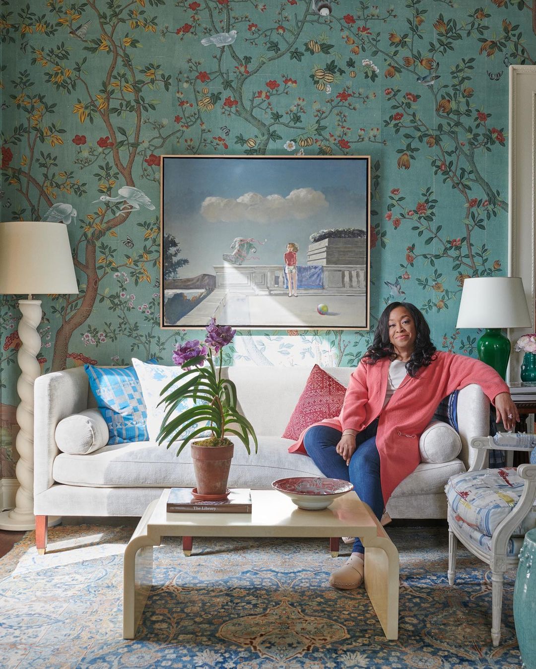 Woman in pink jacket sitting on a white sofa in a room with blue floral wallpaper and a framed landscape painting