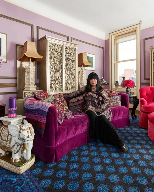 Adult female seated on a purple velvet couch in a room with lavender walls, ornate wooden cabinets, and patterned rug.