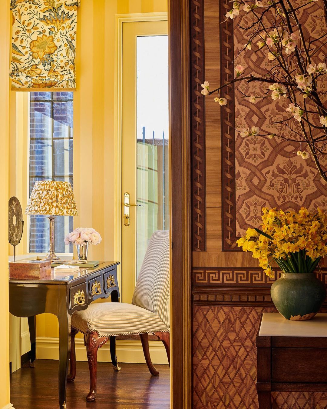 View through a doorway reveals an interior with a desk, chair, floral wallpaper, and a vase of daffodils, creating a warm ambiance.