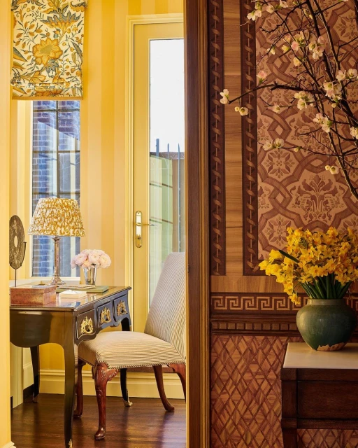 View through a doorway reveals an interior with a desk, chair, floral wallpaper, and a vase of daffodils, creating a warm ambiance.