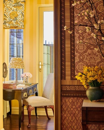 View through a doorway reveals an interior with a desk, chair, floral wallpaper, and a vase of daffodils, creating a warm ambiance.