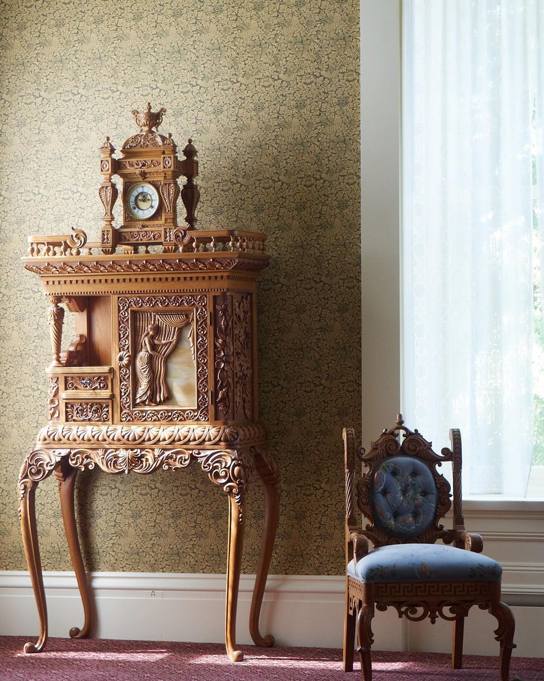 Elaborate antique wooden table with clock and carving details next to ornate wooden chair in front of textured wallpaper indoors.