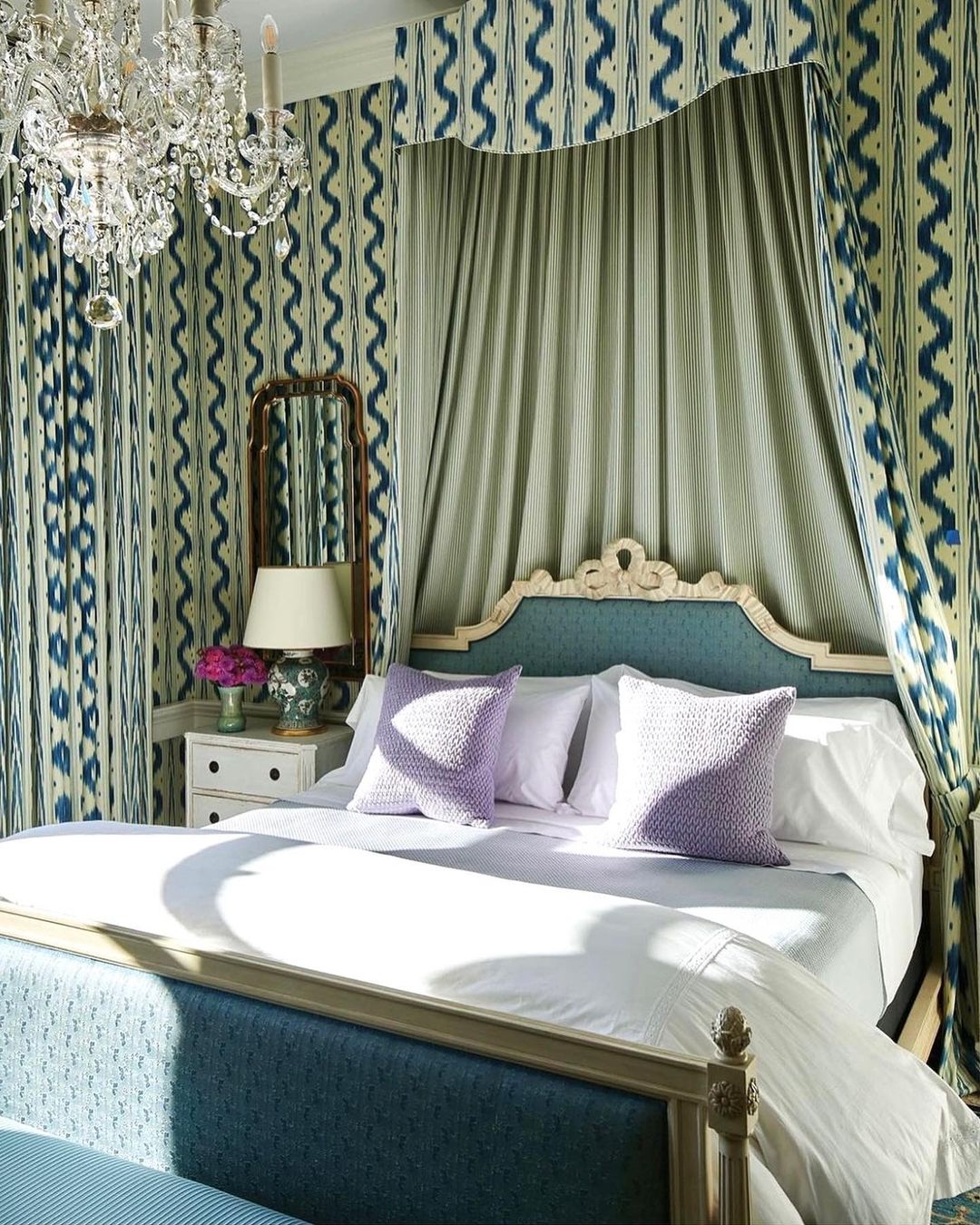 Bedroom interior featuring a bed with white linens, teal headboard, lavender pillows, curtains with blue pattern, and a crystal chandelier.