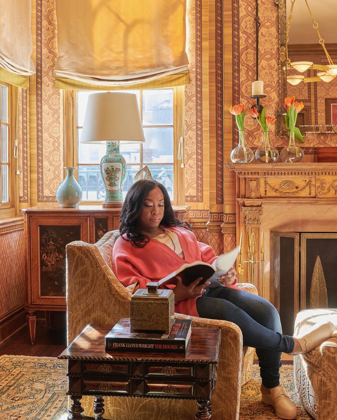 A woman with dark hair sits in a tan armchair in a patterned living room, reading a book. The room features a fireplace and large windows.