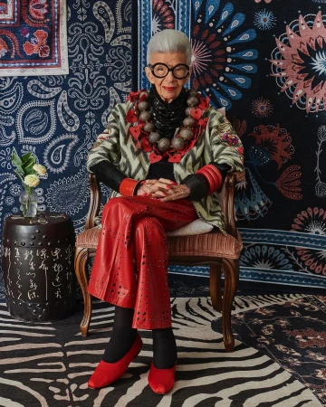 Elderly woman with white hair, wearing glasses and layered jewelry, sits on an ornate chair in front of colorful patterned rugs.