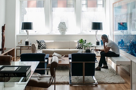 Interior view of a living room featuring a man on his cellphone, cream sofa, black chairs, glass table, artwork, and natural light.