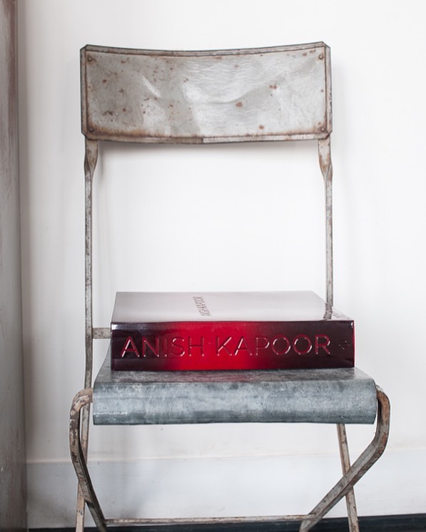A red art book titled 'Anish Kapoor' is displayed on a weathered, antique metal folding chair against a white wall in a neutral setting.