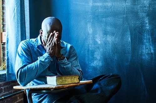 Man sitting at a desk with his hands over his face in an indoor setting next to a window and in front of a blue wall.