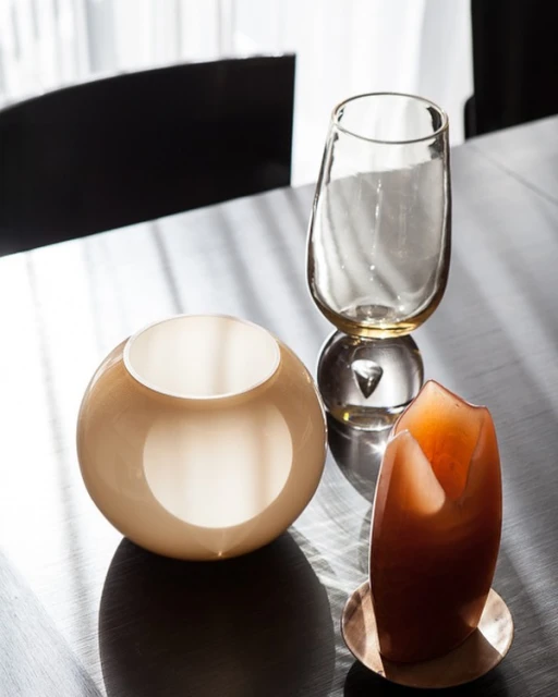 Still life of three decorative glass objects in warm neutral tones with long shadows on a dark tabletop surface.