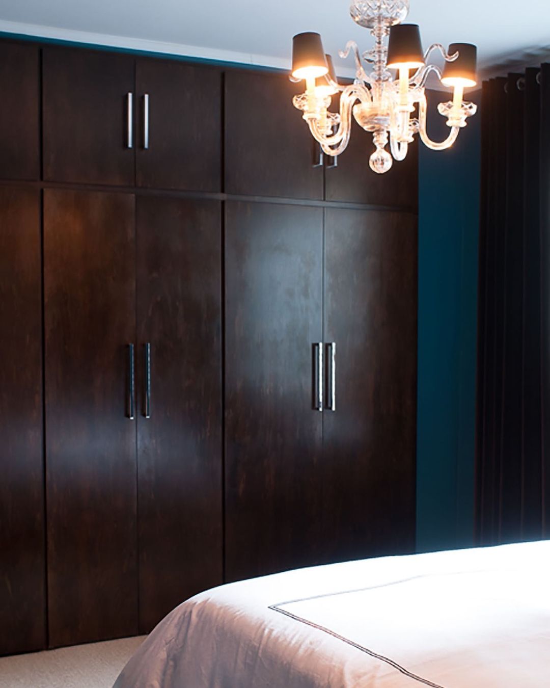 Bedroom interior with a dark wood closet, a crystal chandelier with black shades, and a white bed in a dimly lit room.