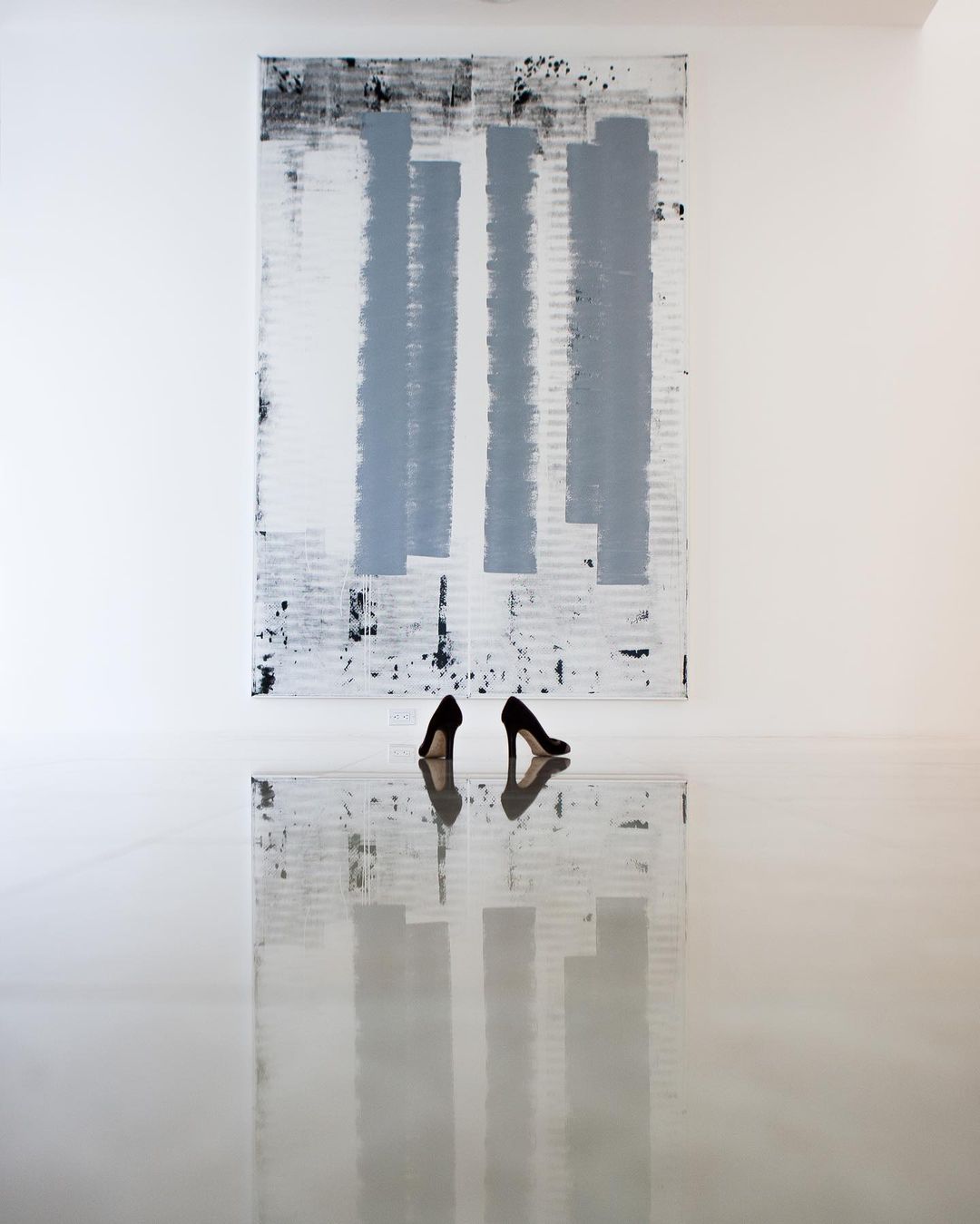 Abstract art display: black high heels mirroring on a reflective white floor in front of a black, grey, and white painted canvas.