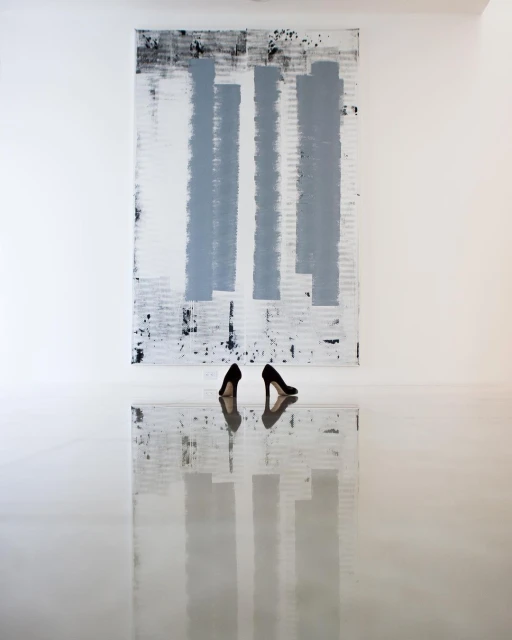 Abstract art display: black high heels mirroring on a reflective white floor in front of a black, grey, and white painted canvas.