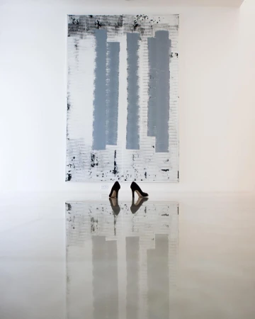 Abstract art display: black high heels mirroring on a reflective white floor in front of a black, grey, and white painted canvas.