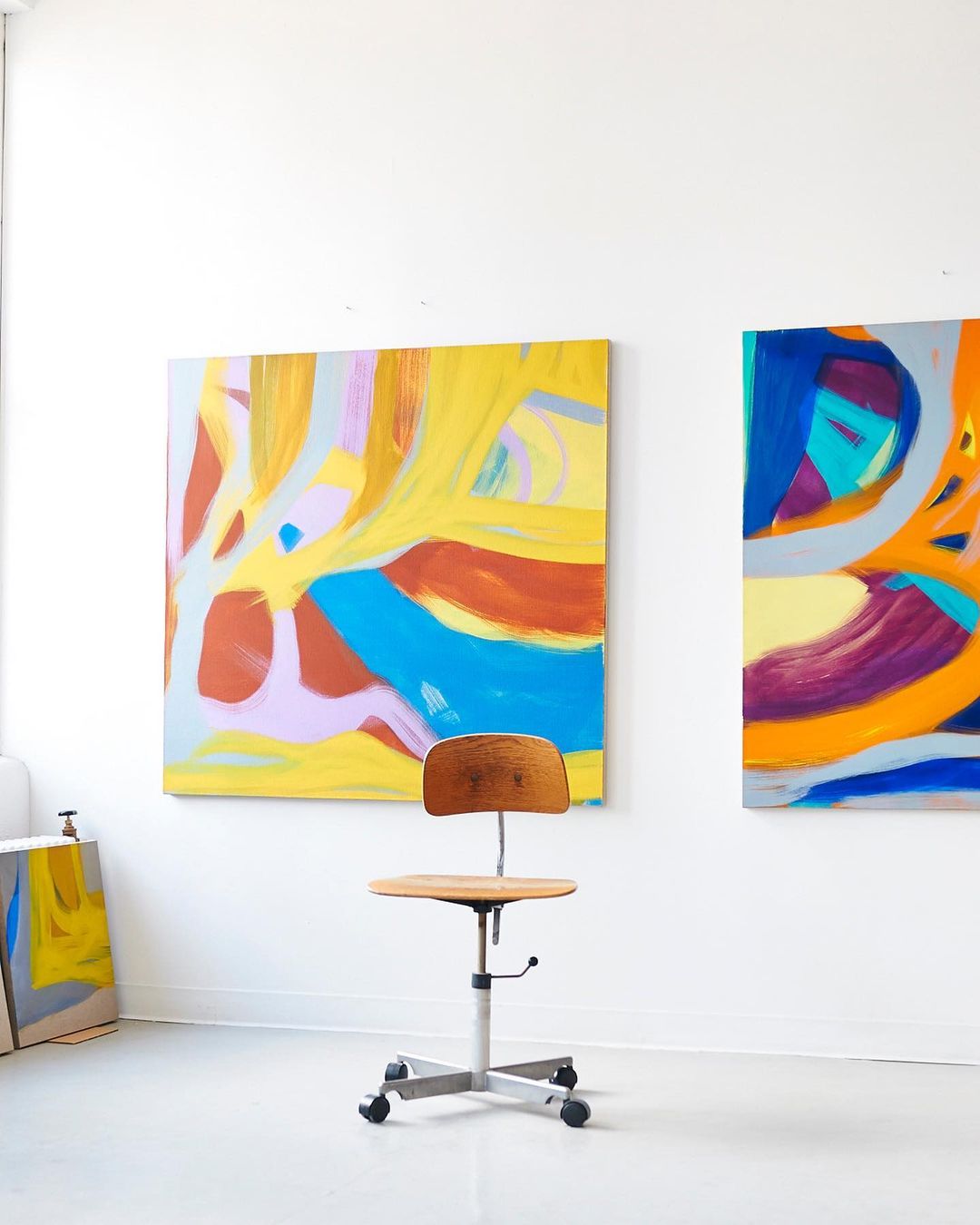 Colorful abstract paintings adorn a white-walled artist's studio, complemented by a wooden chair, creating a vibrant and creative space.