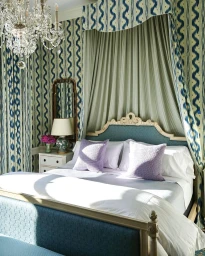 Luxurious traditional bedroom interior with blue patterned wallpaper, elaborate canopy bed, crystal chandelier, and purple pillows